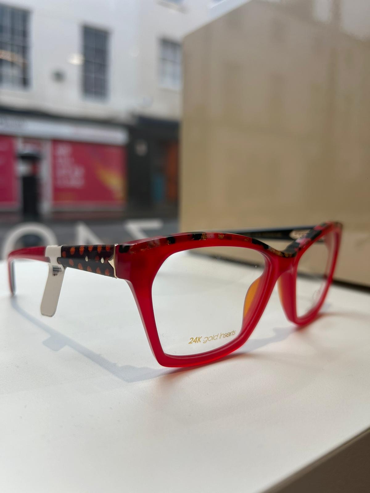 Red and tortoiseshell CCS by Coco Song eyeglasses with 24K gold inserts, displayed on a white surface near a shop window with a blurred street view.