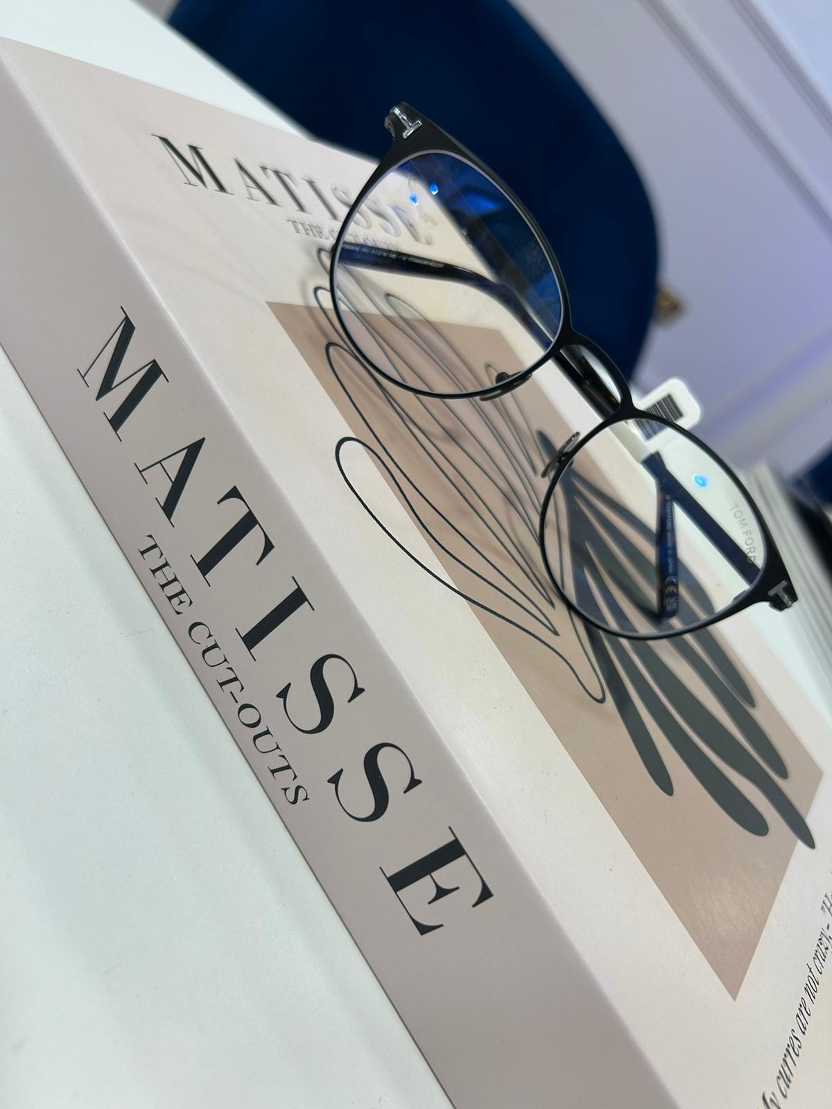 Slim round Tom Ford metal eyeglasses resting on a pink “Matisse: The Cut-Outs” book, with abstract artwork and a navy chair in the background.