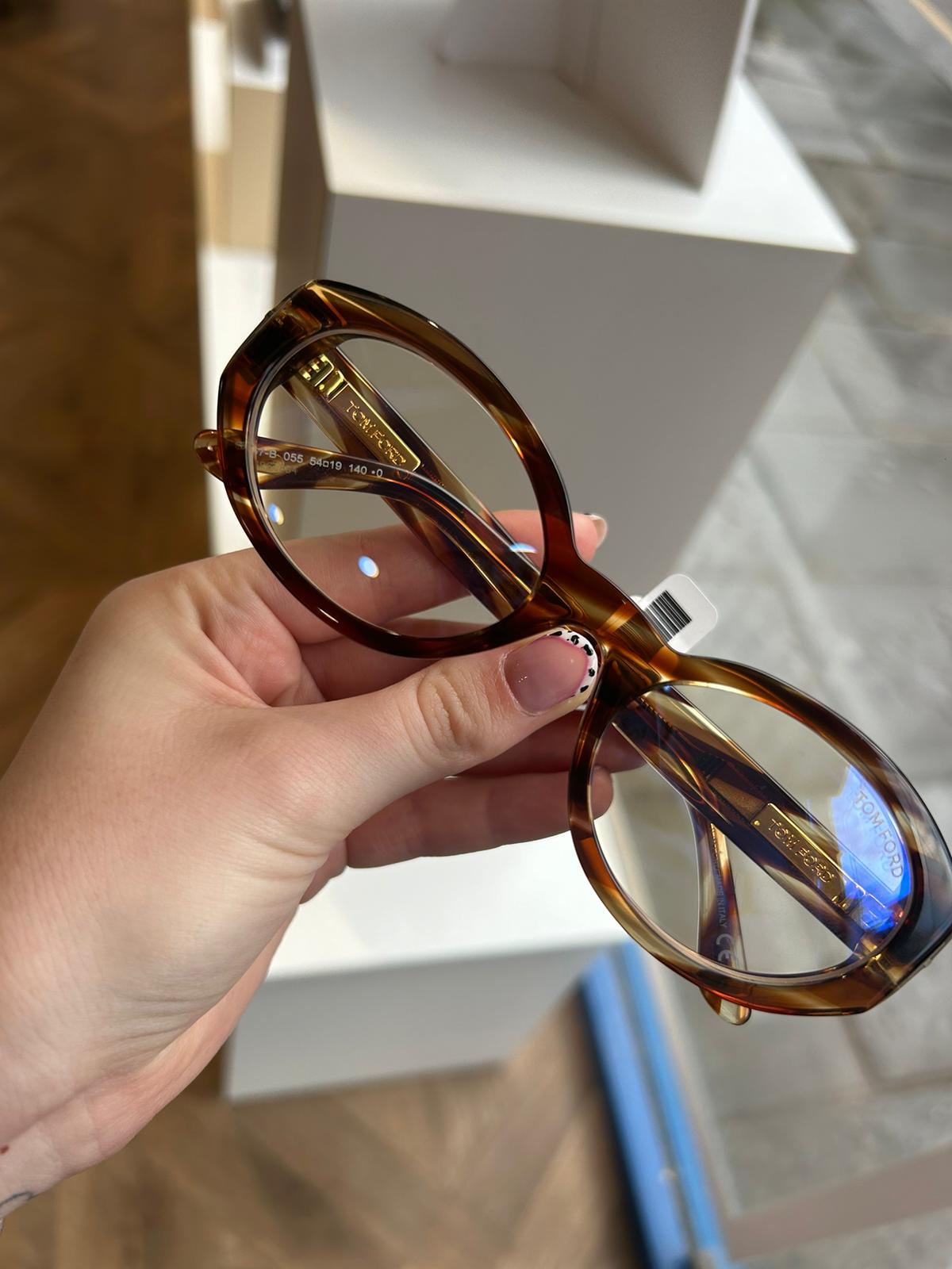 Tortoiseshell Tom Ford oval eyeglasses with gold branding, held in hand inside a modern opticians, with white display stands and a view of the street through the window.