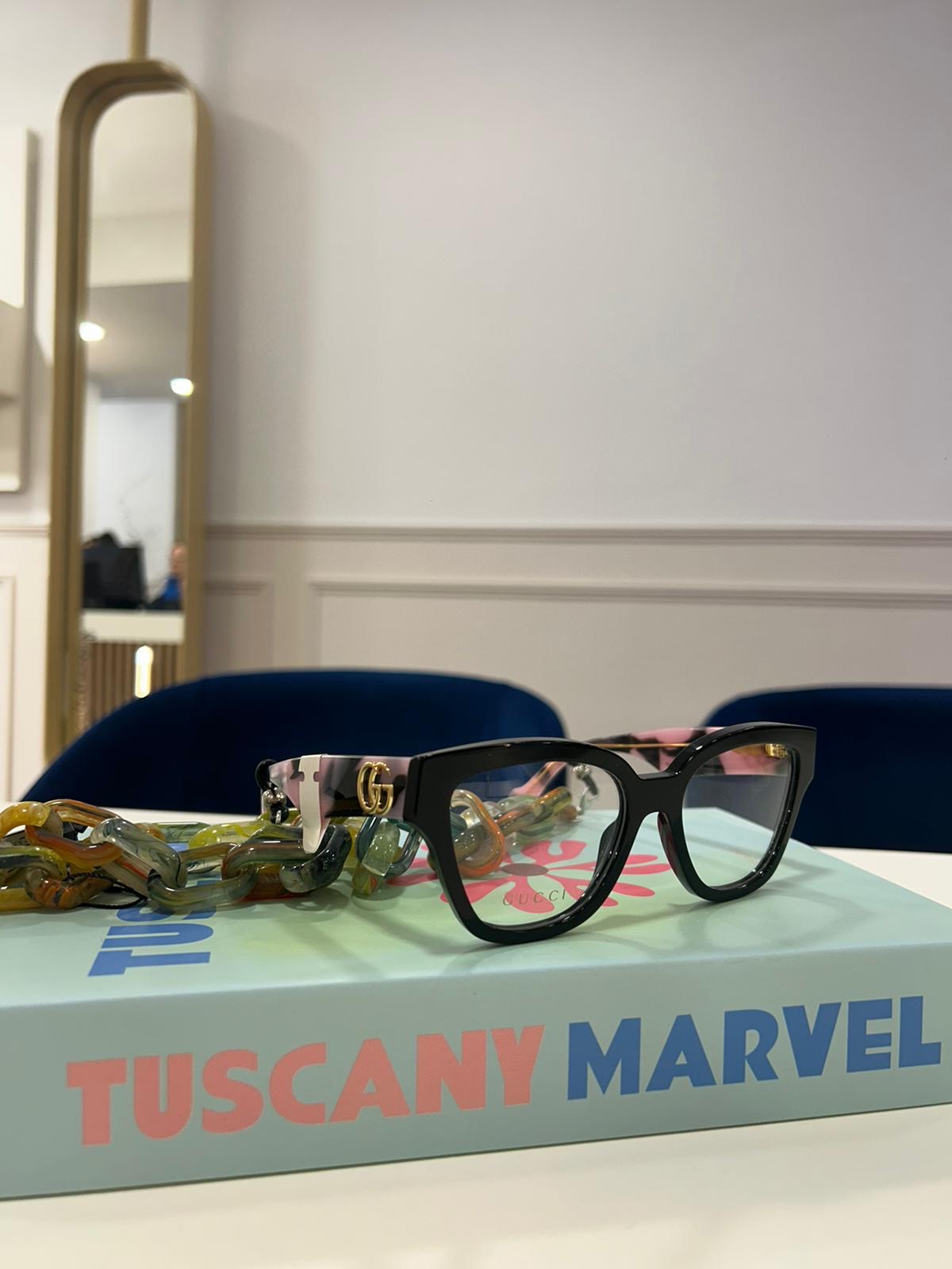 Black Gucci cat-eye glasses with pastel tortoiseshell arms and gold GG detailing, resting on a mint green “Tuscany Marvel” book with a colourful chain attached.