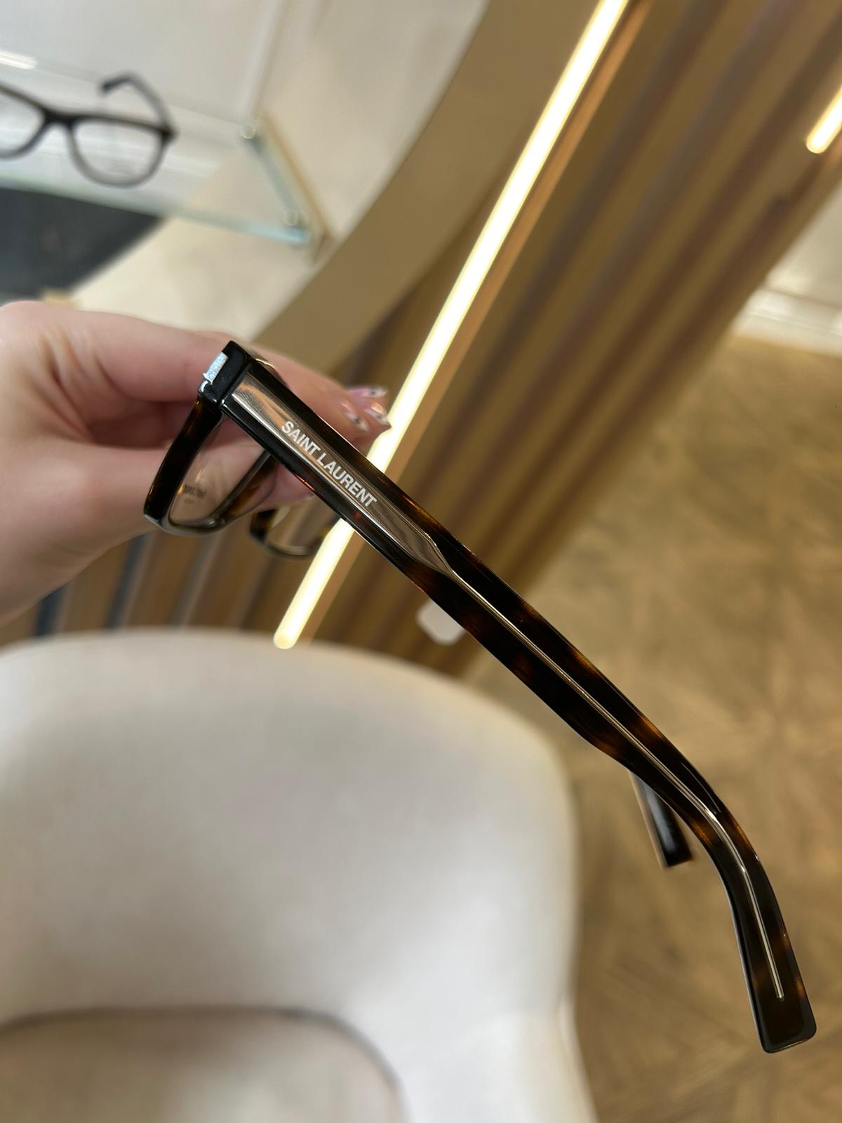 Close-up of tortoiseshell Saint Laurent glasses held in hand, showing the logo on the temple against a stylish optician setting with warm lighting and a cream chair.