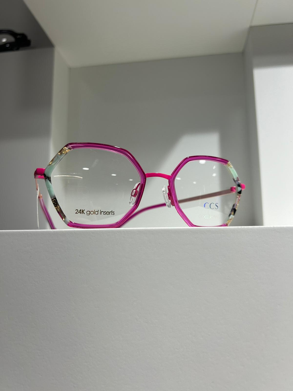 Bright pink geometric CCS by Coco Song eyeglasses with 24K gold inserts, displayed on a white platform against a softly lit background.