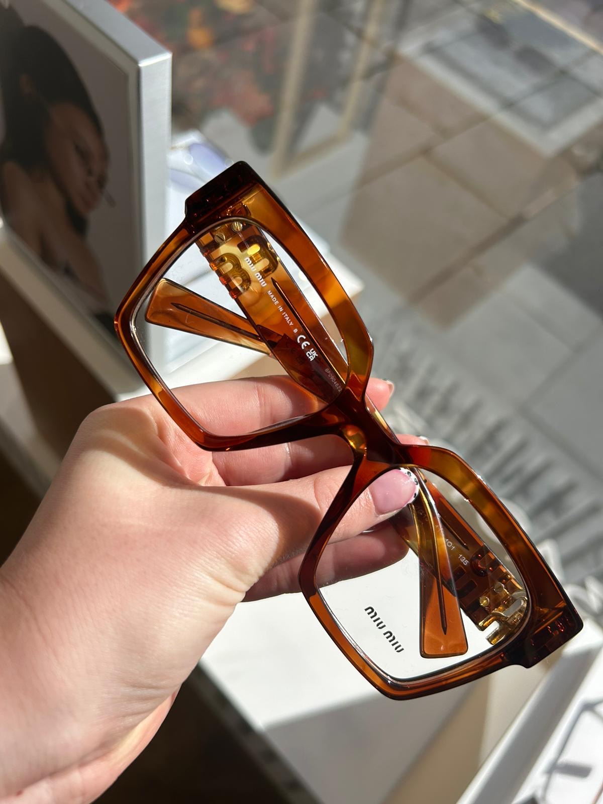 Hand holding oversized tortoiseshell Miu Miu eyeglasses with gold hinge details, near a sunlit window display with a blurred portrait in the background.
