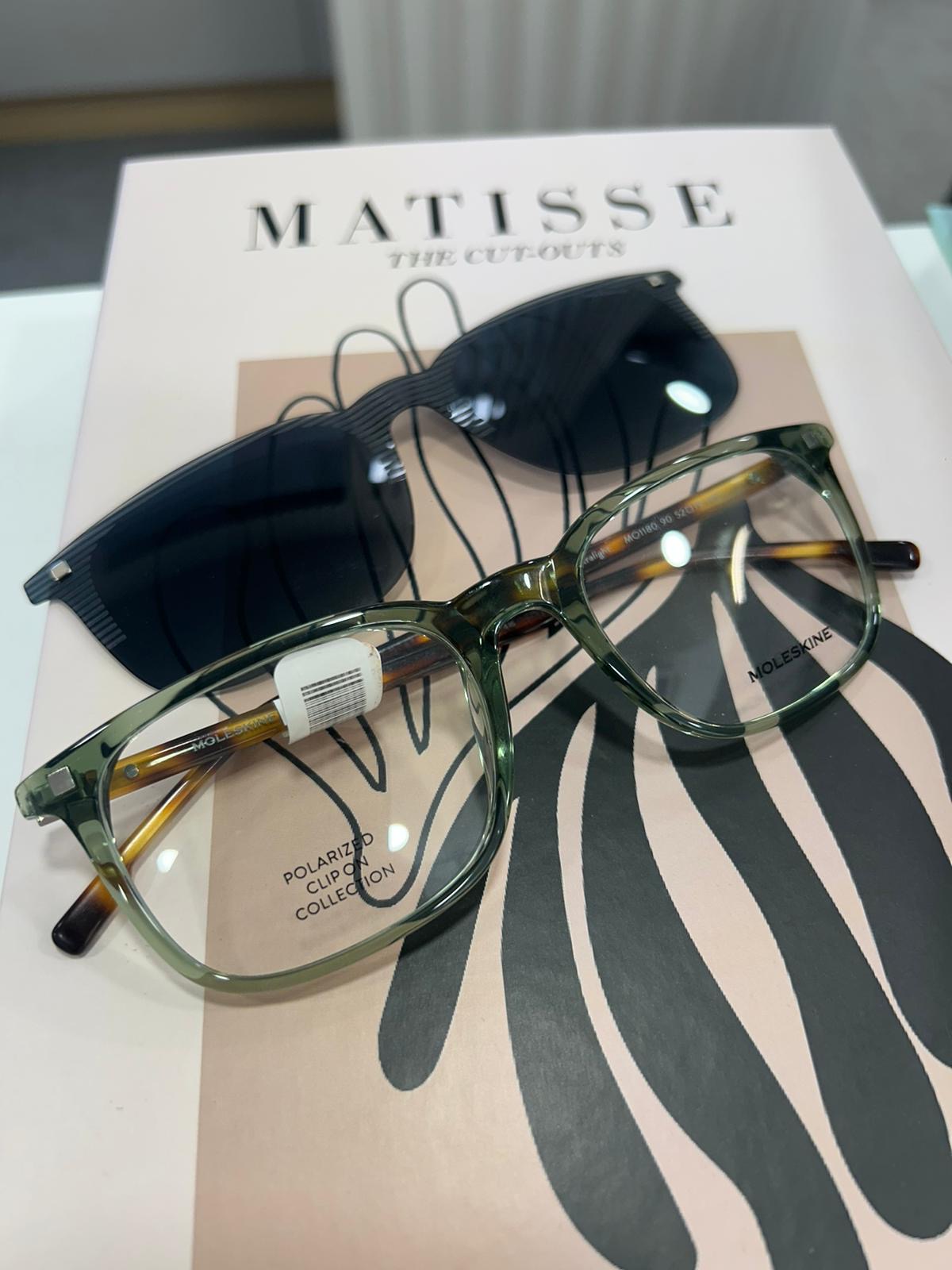 Green Moleskine eyeglasses with a polarized clip-on sunglass attachment, placed on a pink “Matisse: The Cut-Outs” book with abstract artwork.