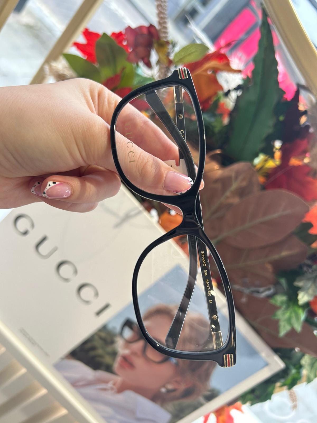 Hand holding black Gucci cat-eye glasses in front of an autumnal window display and Gucci advertisement featuring a woman wearing similar frames.