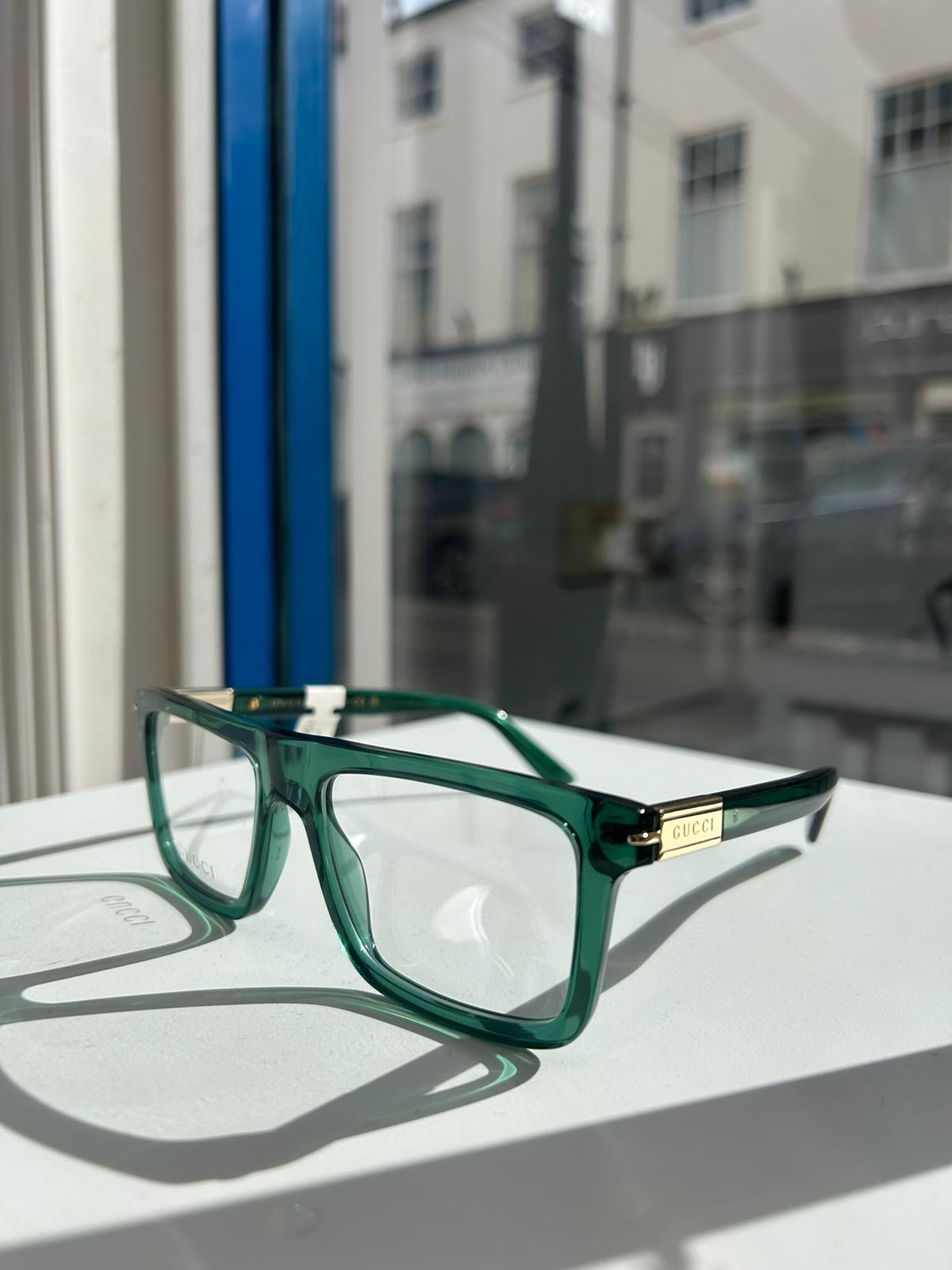 Green square Gucci eyeglasses with gold temple detailing, displayed on a white surface by a shop window with a blurred street view in the background.