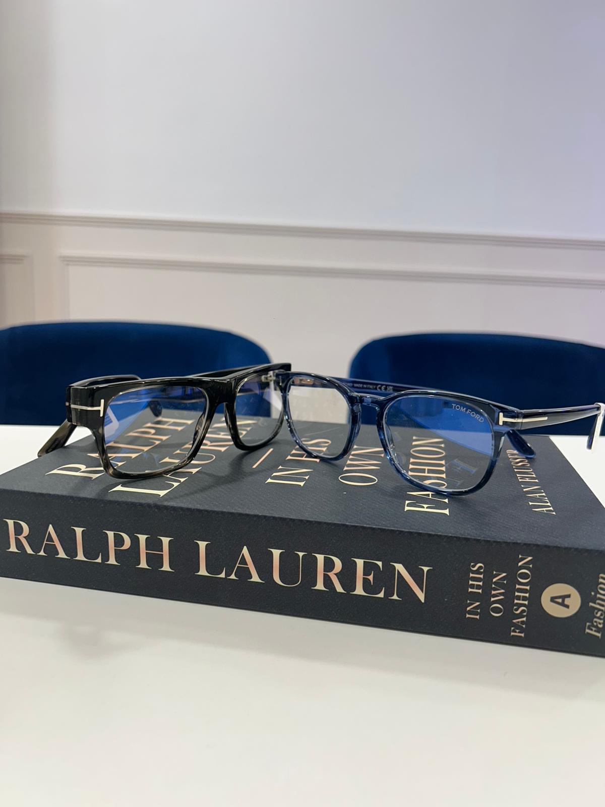 Two pairs of Tom Ford eyeglasses, one black and one translucent navy, resting on a large Ralph Lauren fashion book with blue chairs in the background.