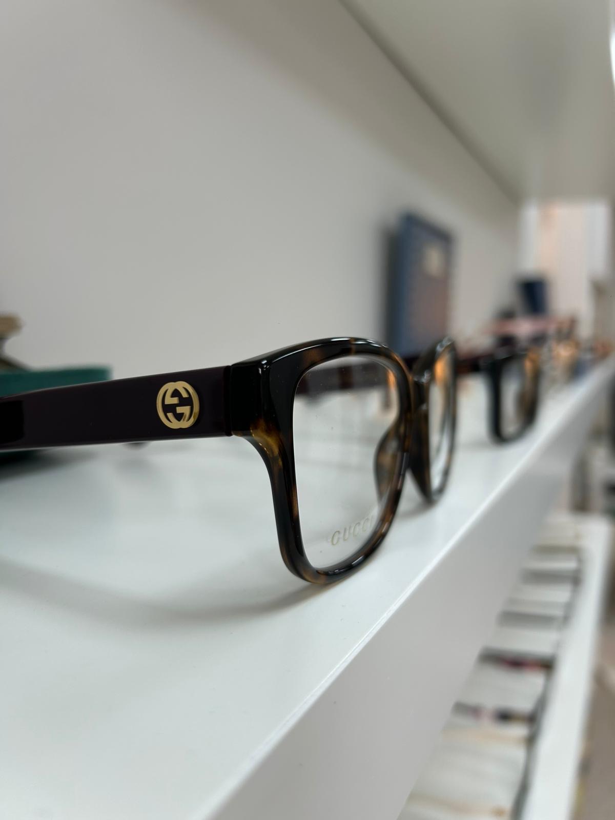 Close-up of Gucci glasses with Havana front, black temples, and gold GG logo, displayed on a white shelf in an optical store.