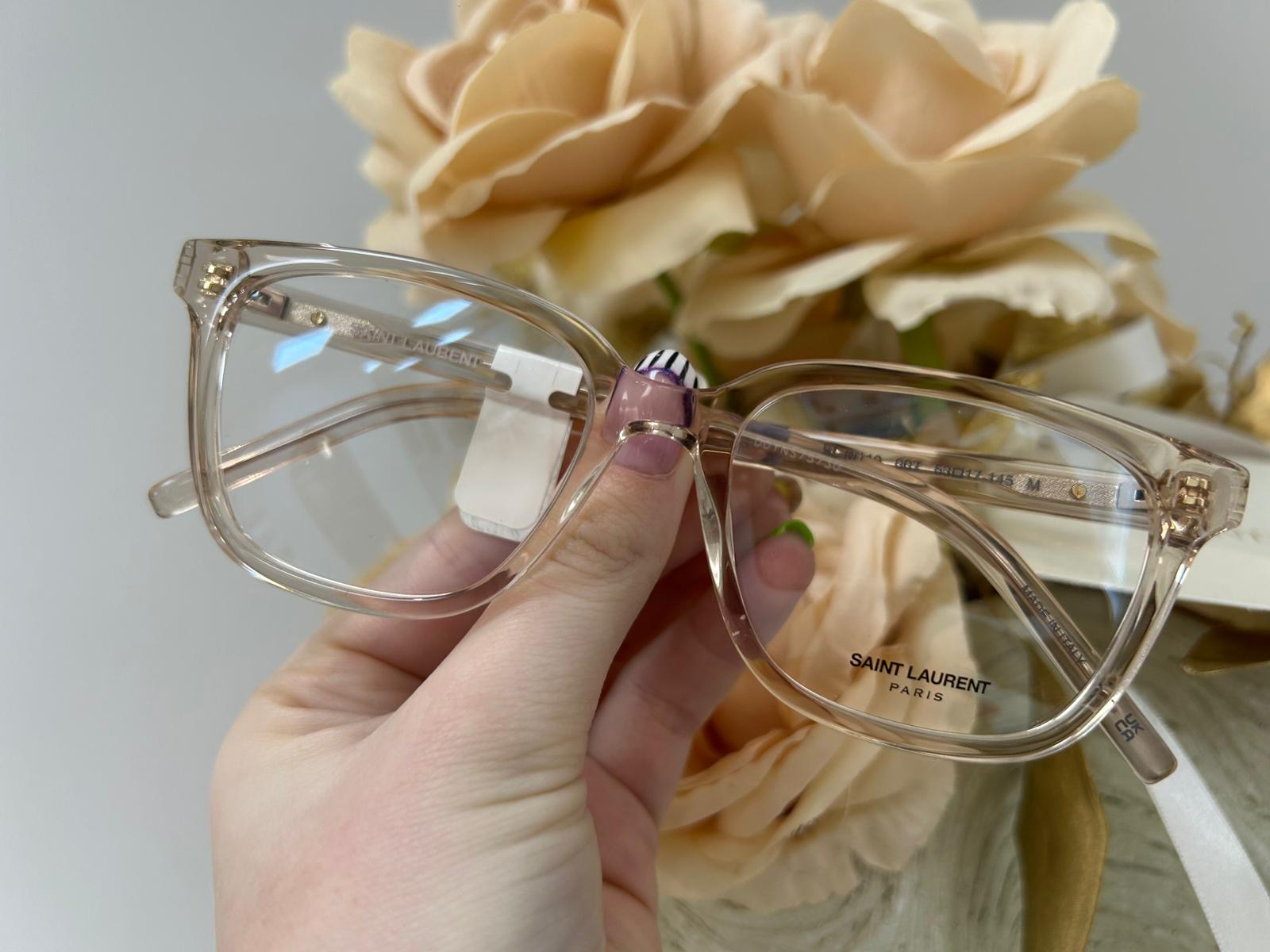 Saint Laurent translucent nude cat-eye glasses with gold accents, held in front of soft peach-coloured roses.