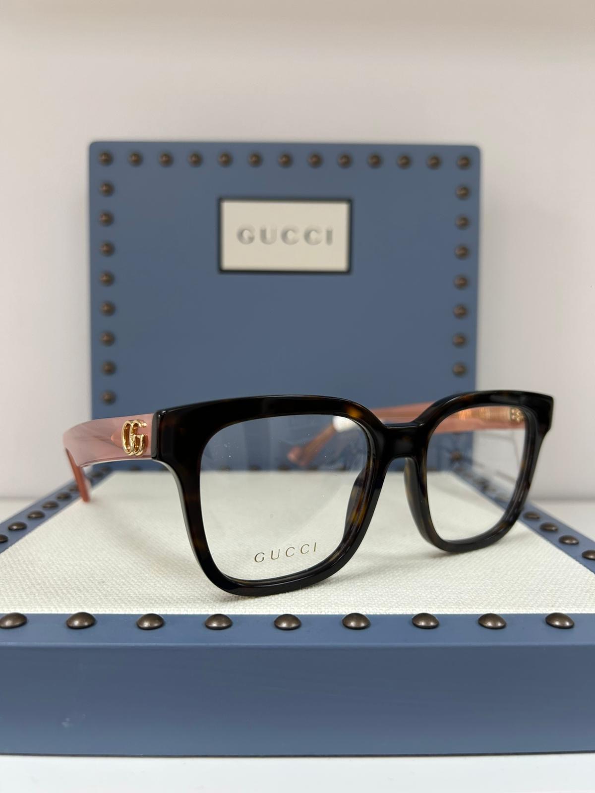 Tortoiseshell and blush Gucci eyeglasses with gold GG detail, displayed on a cream and blue studded presentation box with a Gucci plaque in the background.