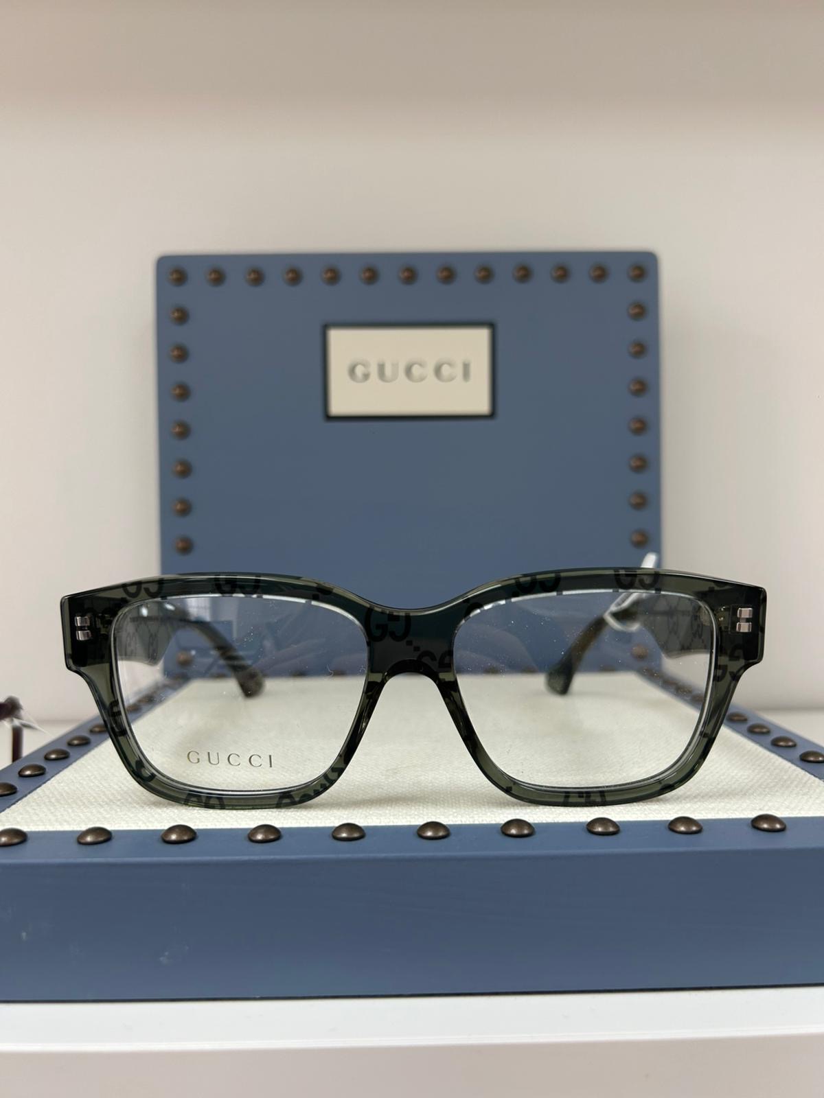 Gucci rectangular glasses in translucent green acetate with tonal GG logo pattern, displayed upright in front of a blue Gucci case with stud detailing.