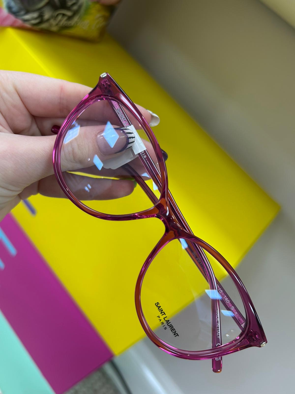 Saint Laurent pink crystal acetate glasses with a rounded frame, held in hand against a bright yellow and pink background.