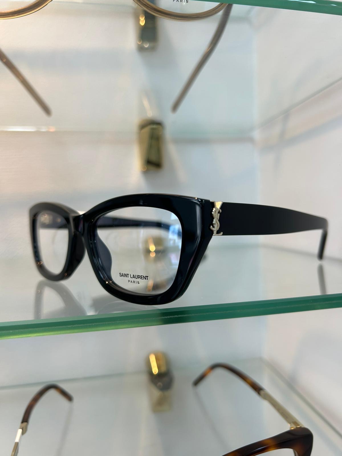 Saint Laurent bold black acetate glasses with oversized square frames and gold YSL logo on the temple, displayed on a glass shelf.
