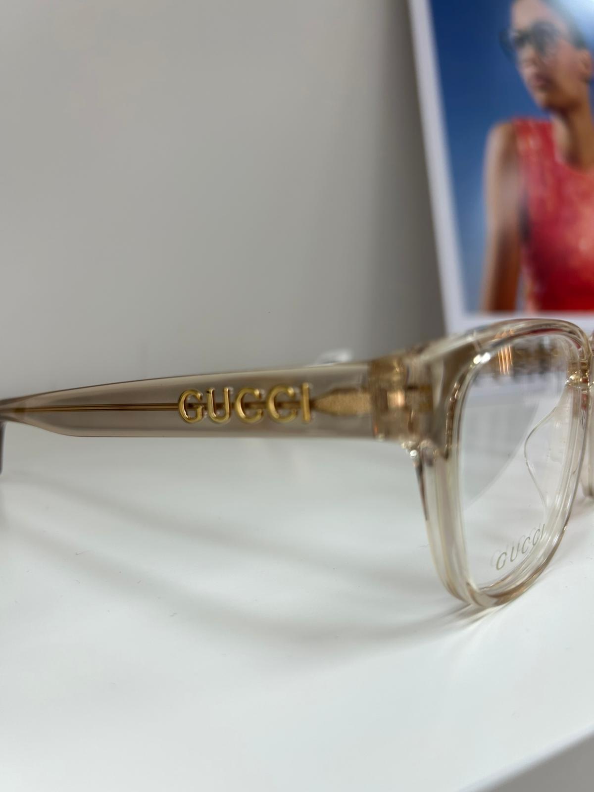 Close-up of translucent champagne Gucci eyeglasses with gold logo on the temple, displayed on a white surface with a blurred fashion poster in the background.
