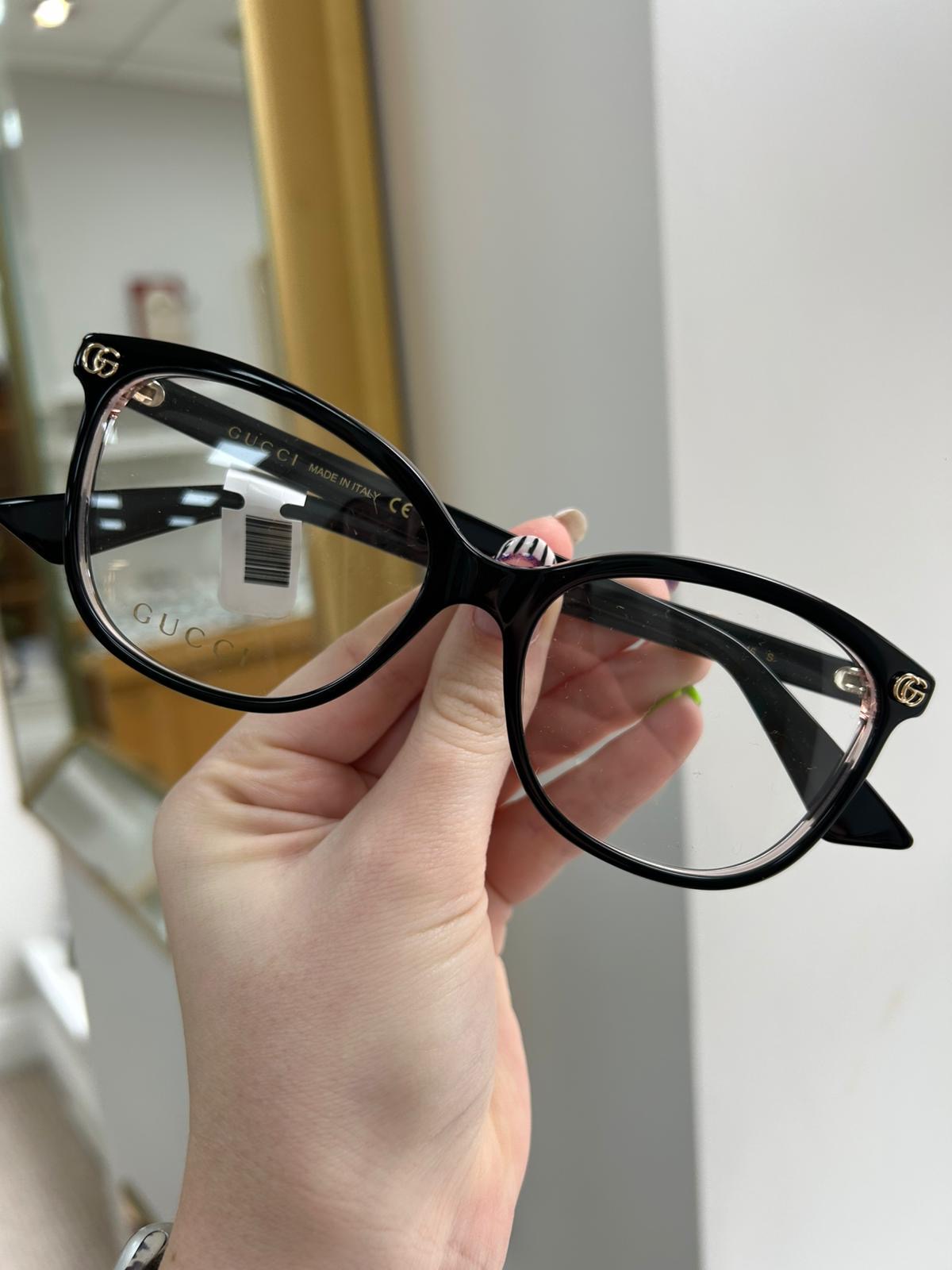 Hand holding a pair of black Gucci cat-eye glasses with gold GG logos on the front corners and pink detailing on the inner rim.