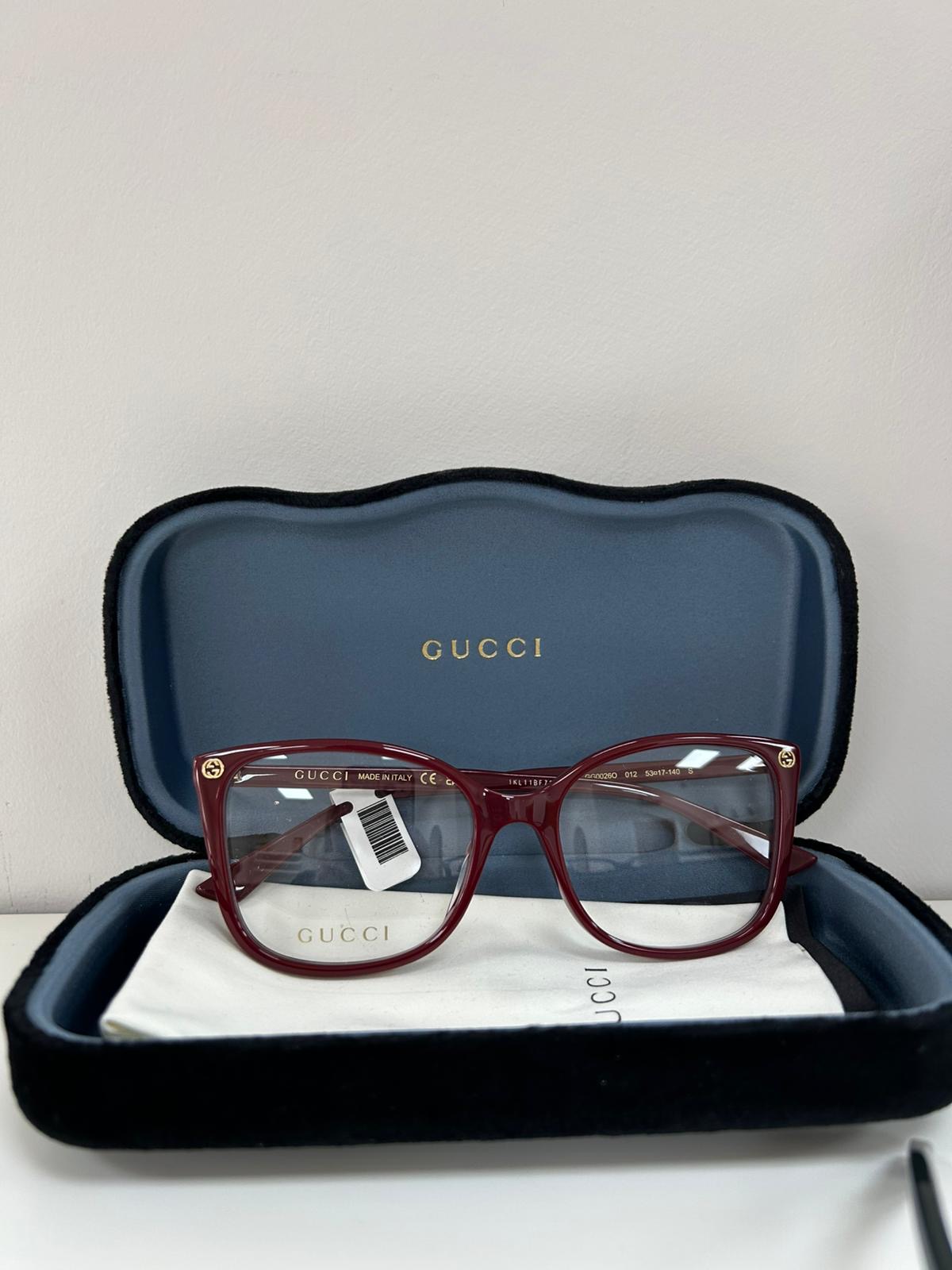 Burgundy Gucci eyeglasses with gold temple accents, displayed inside an open blue velvet Gucci case with a branded cleaning cloth.