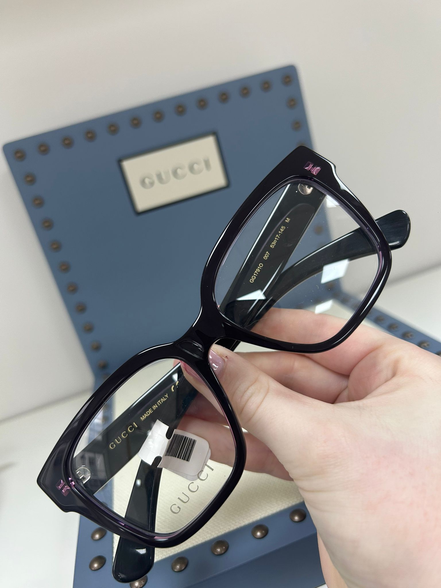 Gucci deep purple square glasses held in front of a blue Gucci display box with stud detailing
