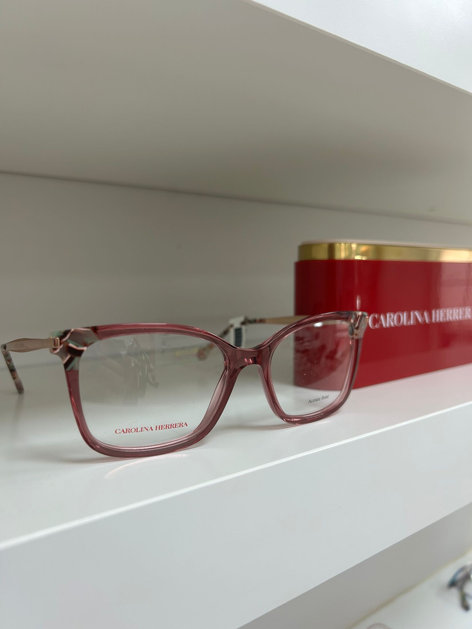 Rose pink Carolina Herrera rectangular glasses with metal temples displayed on a white shelf next to a red and gold branded case.
