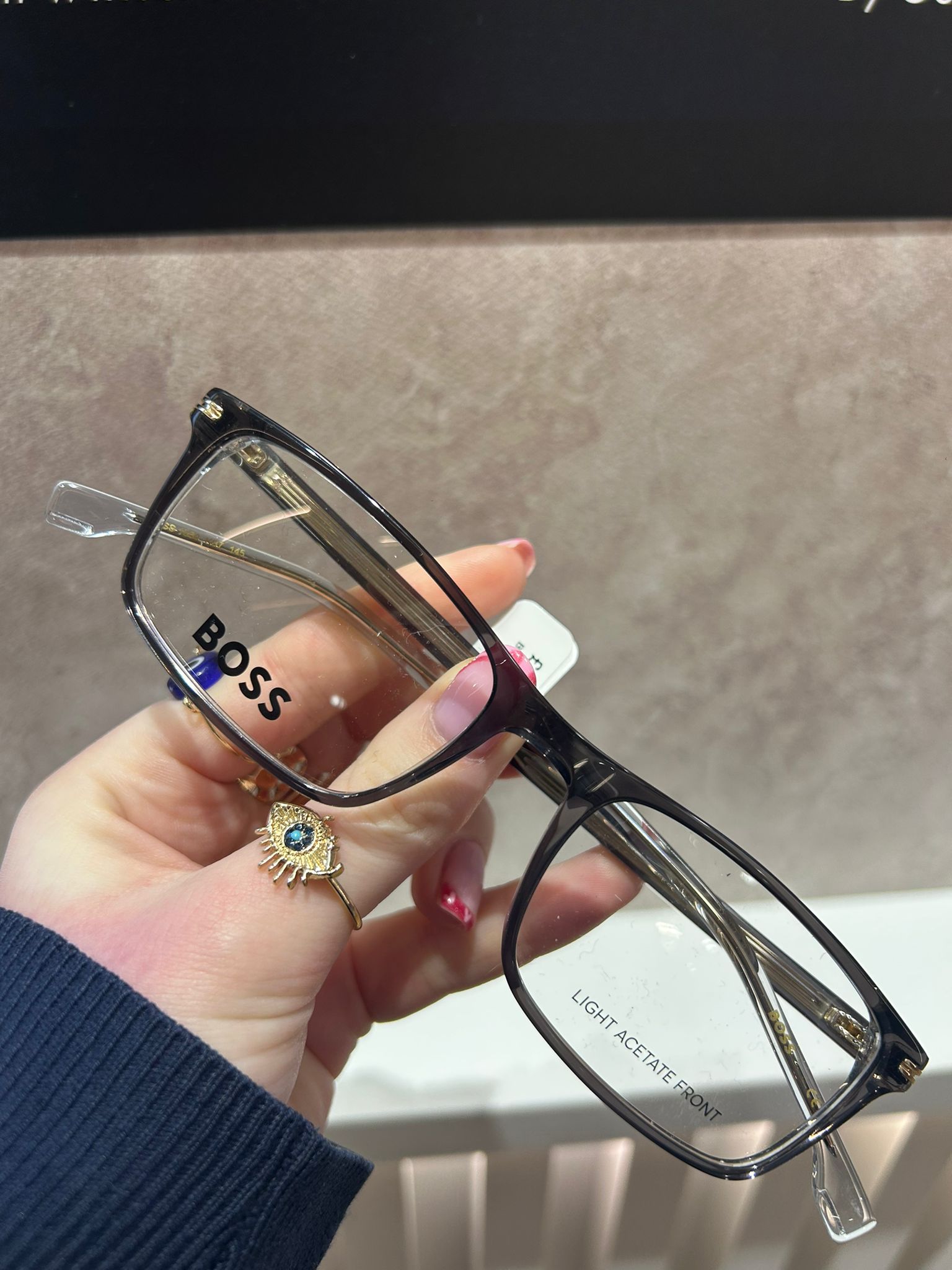 Black rectangular BOSS glasses with clear arms and gold detailing, held in hand against a soft beige background.