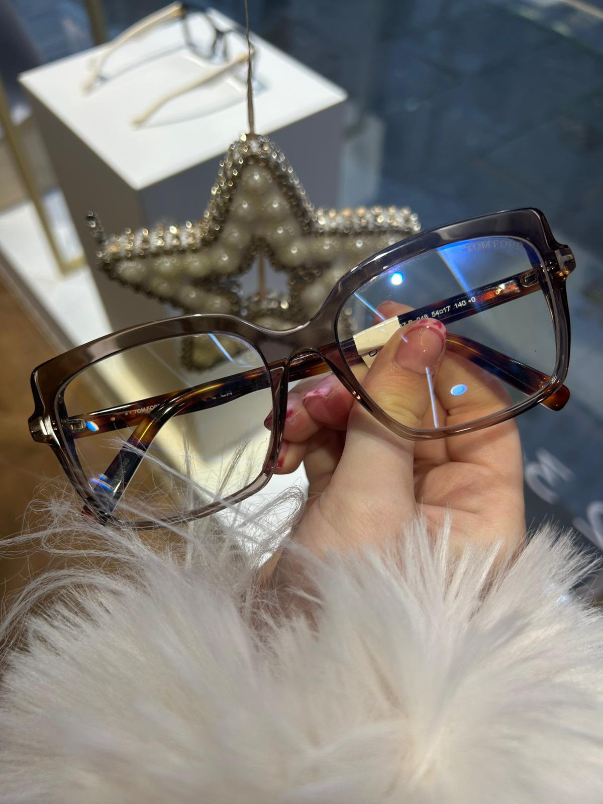 Tom Ford oversized square glasses with gradient brown acetate frame and gold 'T' detailing, held in hand near a shop window display with a pearl-decorated star.