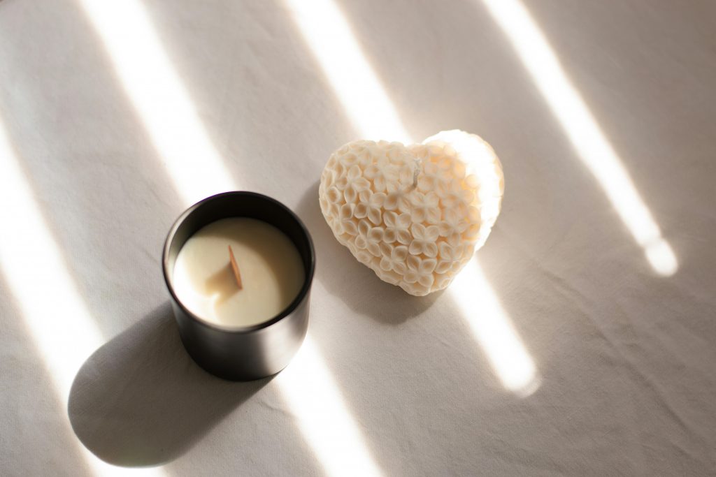 Candle and white heart laying face up against a white background, with strips of sunlight shining on them