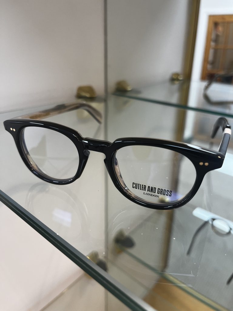 Cutler and Gross round acetate glasses in a dark tortoiseshell finish displayed on a glass shelf, showcasing the brand's distinctive handcrafted design.