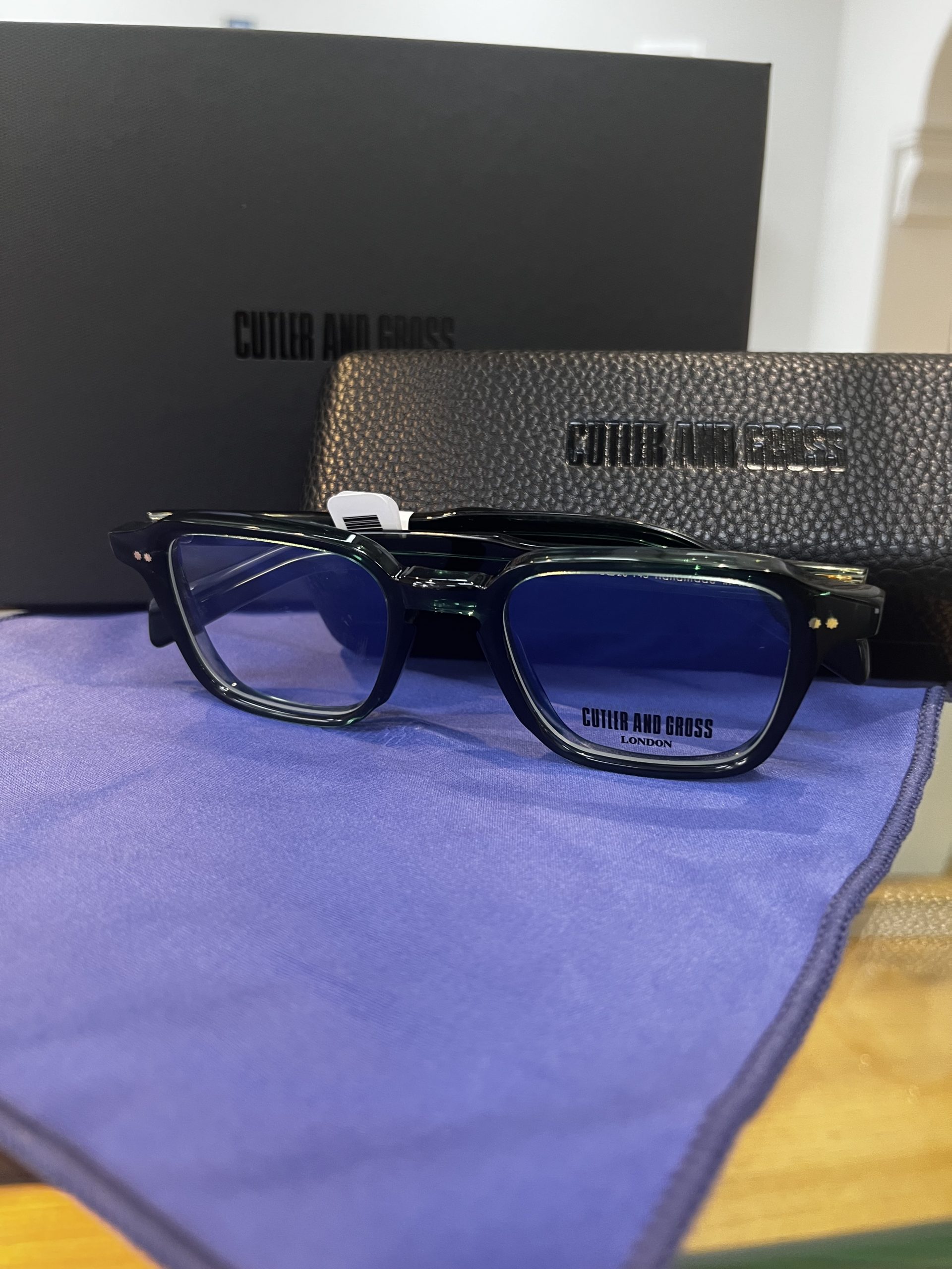 Dark green Cutler and Gross rectangular acetate glasses with blue-tinted lenses displayed on a branded microfibre cloth alongside the presentation case and box.
