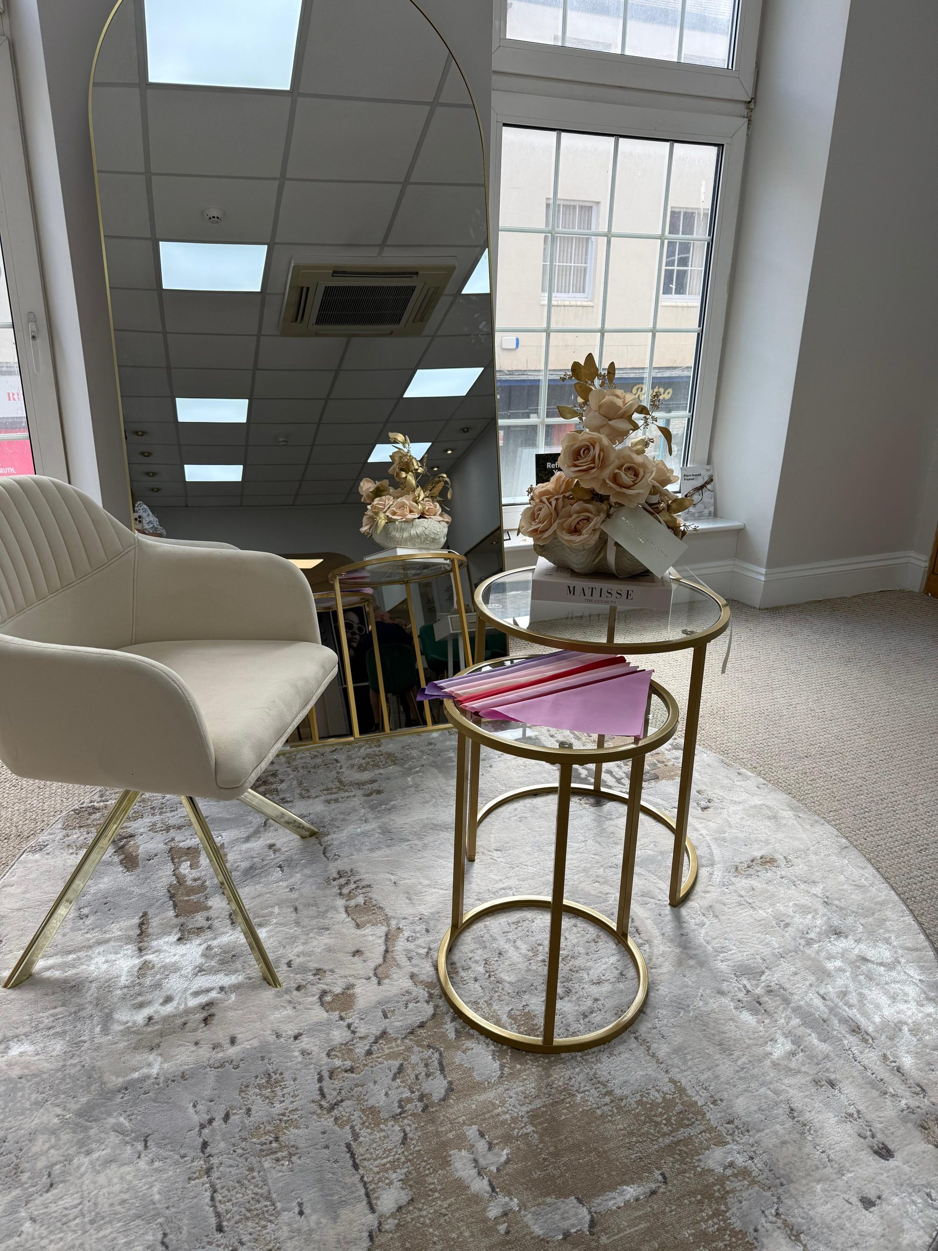 Elegant eyewear styling area inside Edwards & Walker Opticians Doncaster, featuring a cream upholstered chair, gold-framed nesting tables with blush flowers and fabric colour swatches, set beside a large arched mirror and floor-to-ceiling windows.