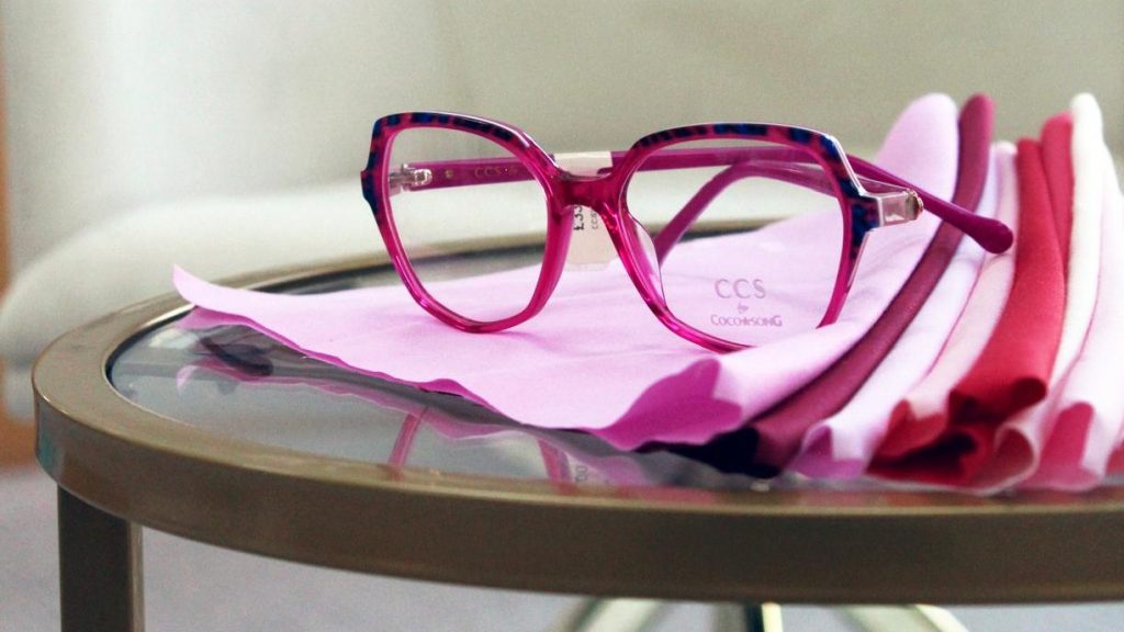 CCS by Coco Song translucent pink geometric glasses with blue tortoiseshell detailing, displayed on folded pink fabric swatches on a glass table in a contemporary opticians in Doncaster