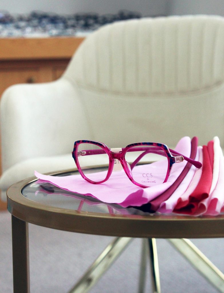 CCS by Coco Song translucent pink geometric glasses with blue tortoiseshell detailing, displayed on folded pink fabric swatches on a glass table in a contemporary optician showroom.