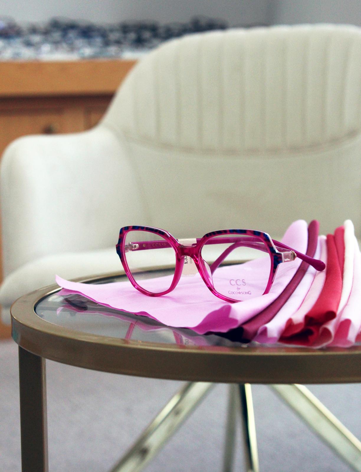 CCS by Coco Song translucent pink geometric glasses with blue tortoiseshell detailing, displayed on folded pink fabric swatches on a glass table in a contemporary optician showroom.